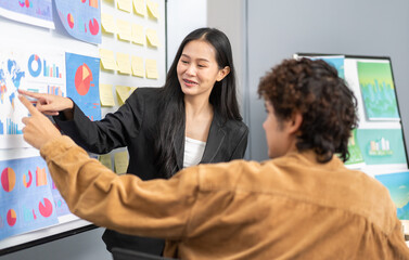 Young businesswoman leading a corporate presentation with global data charts and graphs, explaining business trends and strategies to colleagues during a team meeting in a modern office environment