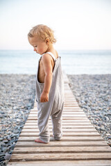 Beautiful toddler in summer grey jumpsuit stands alone on path leading to the seashore on warm day.
