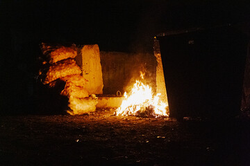 A fire blazing near a rugged stone wall in complete darkness, casting an intense, warm glow....