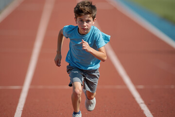 Obraz premium young boy wearing a light blue shirt and gray shorts running energetically on a red track with a blurred background of stadium seats and greenery