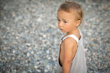 Handsome blond baby with brown eyes looks into distance against backdrop of sea pebbles summer day.