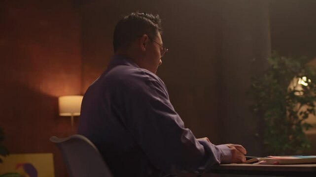 At night time asian businessman with glasses sitting on chair use laptop typing on keyboard. Working. Serious look on screen. 360 degree. Moving around