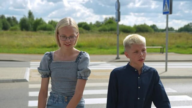 Close up front camera view of brother and sister crossing the street. Happy teen kids chatting while walking outside. Caucasian minors with blond hair, summer outfits.