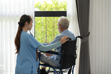 Asian nurse using stethoscope checking on heartbeat examining of senior man sitting on wheelchair....