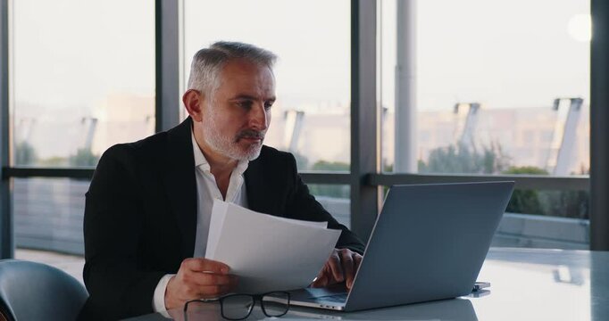 An older, serious gray-haired businessman in a suit works on his laptop and documents in a large modern office. Ideal for showcasing experience, professionalism, and decision-making in business.