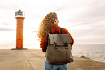 Young woman enjoying a breezy day near a vibrant red lighthouse.  Autumn sunset at a background. Travel, weekend, relax and lifestyle concept.