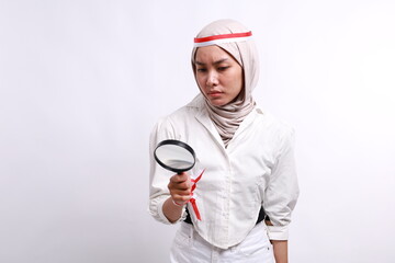 Curious Asian muslim woman wearing red white flag ribbon, standing while checking something using a magnifying glass, isolated on a white, celebrate Indonesia independence day