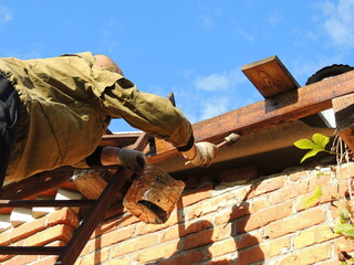 man on ladder with dark liquid in plastic jar and brush in hands soaking beam on house roof, protection of wooden material during construction by oiling