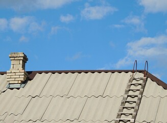 Fototapeta premium chimney brick pipe and wooden ladder on gray corrugated slate roof of private rural house under clear blue sky, top of rustic building with stairs and kitchen pipe for smoke outlet as country texture