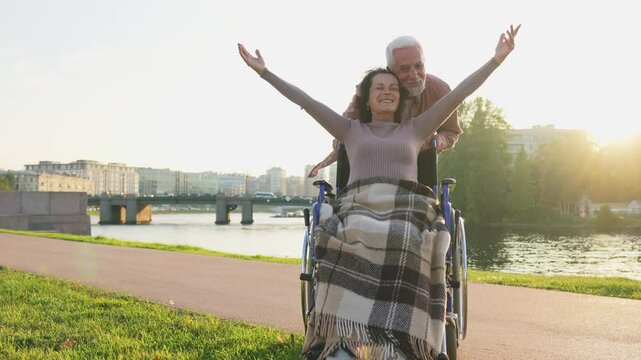 Senior woman in wheelchair walking with caregiver old man on road. Family couple man husband supporting taking care of paralyzed woman wife in chair for people with disability outdoor. Rehabilitation
