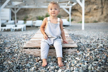 A beautiful blond baby with brown eyes, tanned, sits on a wooden path leading to the seashore made of pebbles on a warm summer day.