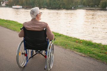 Sad senior man sitting in wheelchair walking on road in city park outdoor, back view. Old paralyzed man in chair for people with disability feel depressed. Lonely elderly sick man sits on wheelchair