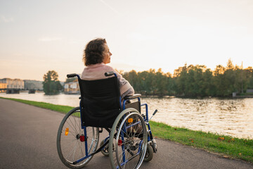 Naklejka premium Sad senior woman sitting in wheelchair walking on road in city outdoor, back view. Old paralyzed lady in chair for people with disability feel depressed. Lonely elderly sick woman sits on wheelchair