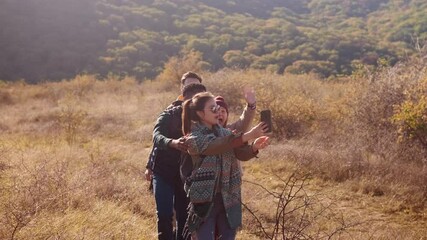 Smiling group of friends having fun while hiking together on hill at sunset and making video blog with smart phone.