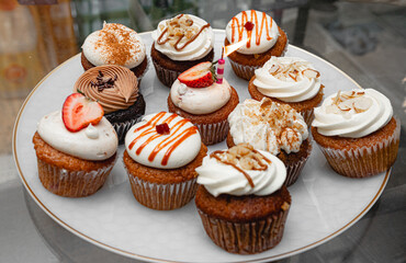 A decorative plate with an assortment of cupcakes topped with frosting, fruit slices, and drizzles, showcasing various flavors and designs for a dessert table.