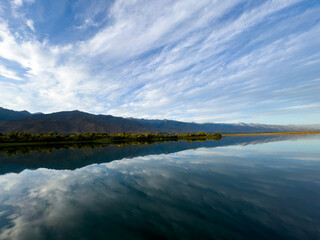 Fototapeta premium The calm water of a mountain lake reflects the blue sky and white clouds. The green shoreline frames the lake, creating a sense of harmony and tranquility.