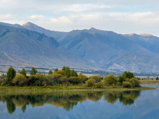 The calm water of a mountain lake reflects the blue sky and white clouds. The green shoreline frames the lake, creating a sense of harmony and tranquility.