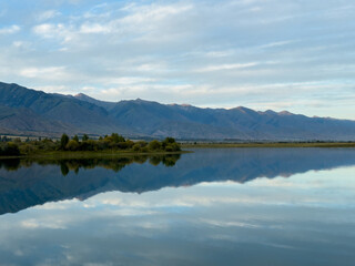 The calm water of a mountain lake reflects the blue sky and white clouds. The green shoreline frames the lake, creating a sense of harmony and tranquility.