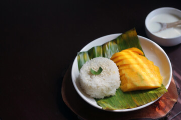Mango sticky rice, a popular Thai dessert. The plate features beautifully sliced, ripe golden mango, a mound of glutinous white rice garnished with sesame seeds and a bowl of coconut milk.