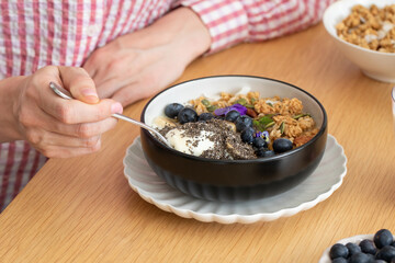 A woman is sitting at a table, eating a healthy breakfast. Granola with Greek yogurt and blueberries