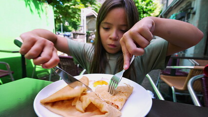 Young girl cutting into a crepe with a knife and fork at an outdoor café, focusing intently on...