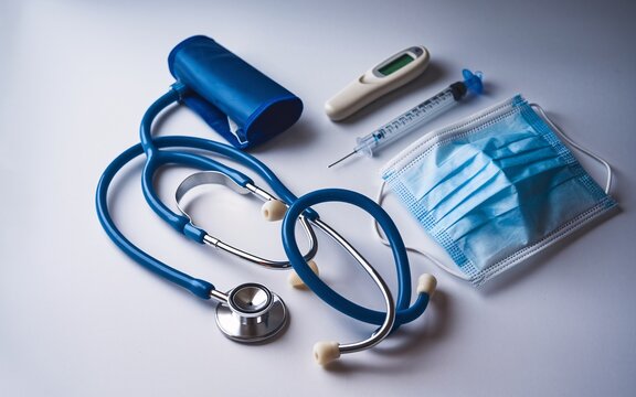 Medical equipment laid out on a white background, including a stethoscope, a blood pressure cuff, a thermometer, a syringe, and a face mask.