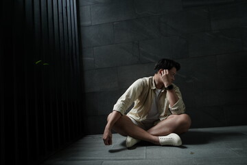 Asian man in shorts sits on the floor in a dark corner outside his house feeling bored, anxious, and lonely.