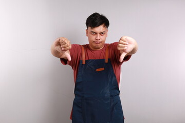 Upset asian man wearing blue apron frowning sulking sad, disappointed, show thumbs-down grimacing,...