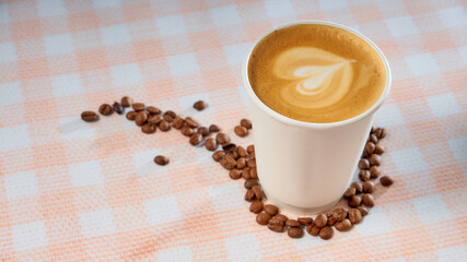 Takeaway coffee paper cup with coffee beans on a checkered background. Milk foam heart