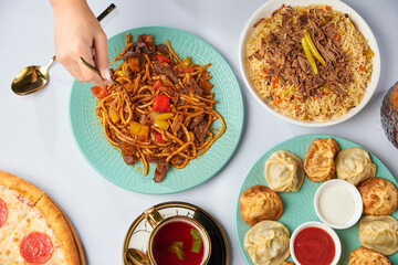 The table is filled with dishes and food. Set of Uzbek pilaf, lagman, manti. Woman's hand with a fork. Food background.