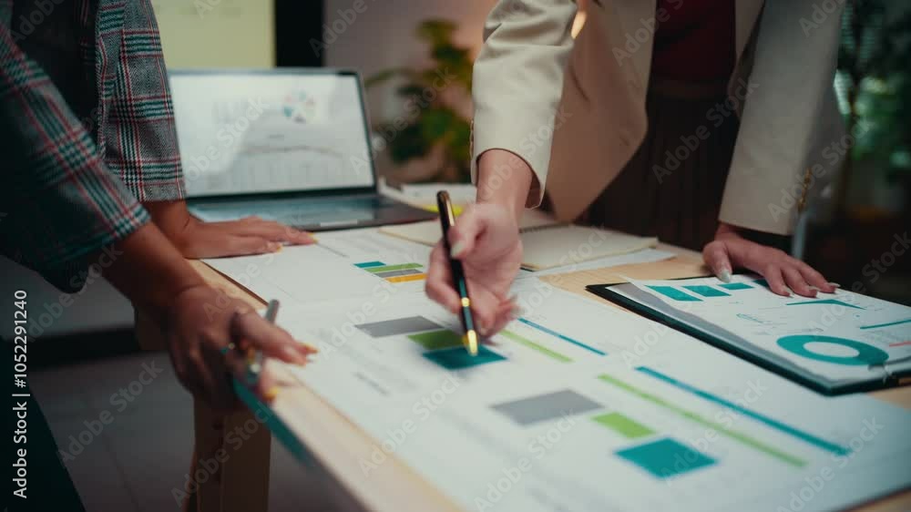 Poster Two businesswomen are collaborating on a project, standing by a table and reviewing financial reports. They are pointing at charts and graphs, discussing data and insights