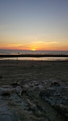 Fototapeta premium This is a beautiful sunset photograph taken on a beach. The sun is low on the horizon, casting warm orange and yellow hues across the sky and reflecting on the water. The scene includes several people