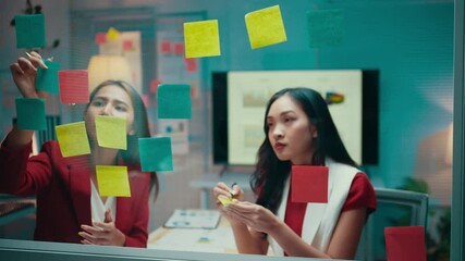 Businesswomen engage in a meeting, writing on sticky notes and placing them on a glass wall. The collaborative atmosphere fosters creativity and strategic planning