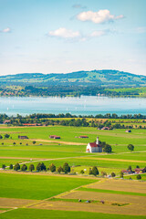 Beautiful landscape with green fields, white church, blue lake, mountains and sky with clouds in the background. Vertical photo