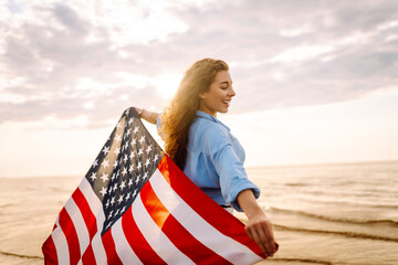 Young woman holding national American flag walking ocean beach. America Independence Day concept....