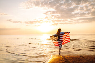 Young woman holding national American flag walking ocean beach. America Independence Day concept....