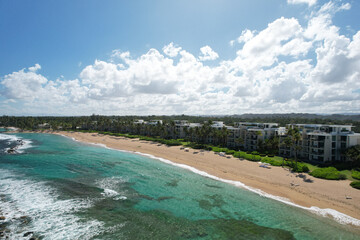 Aerial vantage of the west beach residences at Dorado Beach Resort, Puerto Rico, showing the coastline, sandy beach, palm trees, under a cloudy blue sky.