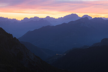 Fototapeta premium Twilight view of the Italian Dolomites from Giau Pass, with a colorful sunset sky illuminating the rugged mountains and a distant village Selva di Cadore in the valley.