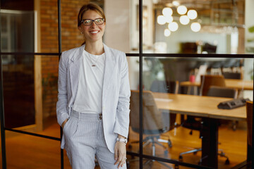 A confident and successful businesswoman dressed in a chic suit, standing proudly in a modern...