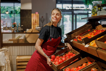 A cheerful woman is happily organizing and arranging ripe tomatoes in a warm and cozy grocery store environment
