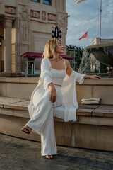 Obraz premium Woman White Outfit Fountain - Sitting on bench near a fountain in an urban setting, a woman in a white outfit poses for a photo.