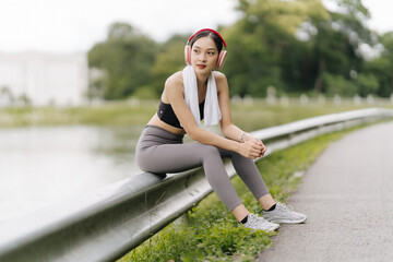 Resting After Workout: A young woman in sportswear sits on a bridge after a run, taking a break to listen to music.  She is wearing headphones and a towel around her neck.