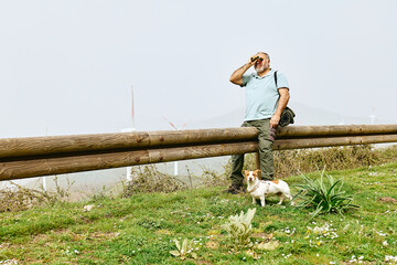 Male tourist using binoculars in front of wind turbine in wind farm with mountain landscape view.