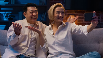 Happy excited multiracial multiethnic couple married diverse Asian man and Caucasian woman talking online video call conference with mobile phone app talk waving hello at night evening on couch sofa