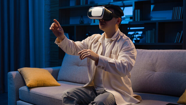 Asian adult man using 3d goggles eyeglasses for playing game at home at night Korean male wear VR glasses helmet play metaverse using virtual reality for business working project at evening at couch