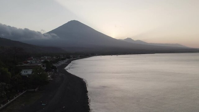 Atardecer en una playa de arena negra y palmeras en Bali d&oacute;nde el sol se pone detr&aacute;s de un volc&aacute;n y nubes bajas. Barco atracado en la orilla y personas en la arena.