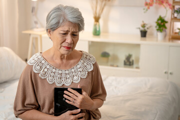 An elderly Asian woman with short white hair is thinking of her loved one, hugging a picture of the person she is missing and crying.