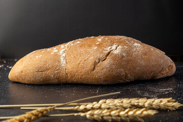 A rustic loaf of bread with unique textures sits alongside wheat stalks, placed creatively on a dark background, ideal for concepts of food artistry or rustic lifestyles.
