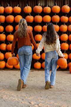 two girls holding hands, seen from behind, holding pumpkins and walking towards pumpkin wall