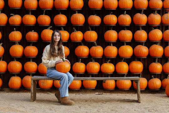 brown haired caucasian girl holding a pumpkin, sitting on a bench in front of a wall of pumpkins